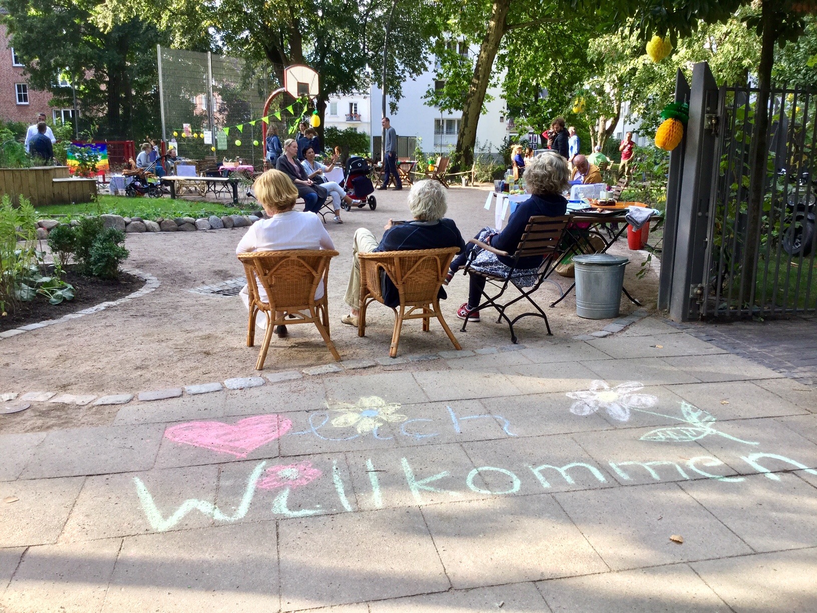 Nachbarschaftsfest, Blick in den Garten von der Lutterothstraße aus. Auf dem Gehweg steht in bunten Buchstaben Willkommen und drei Menschen aus der Gartengruppe sitzen auf Stühlen mit dem Rücken zum Betrachter 